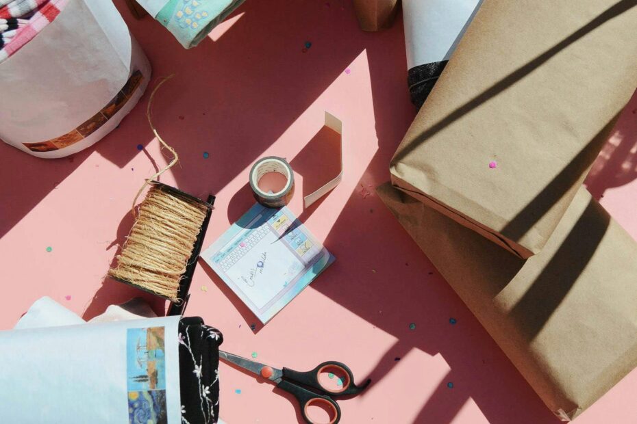 Top view of gift wrapping supplies including paper, scissors, and tape on a pink background.