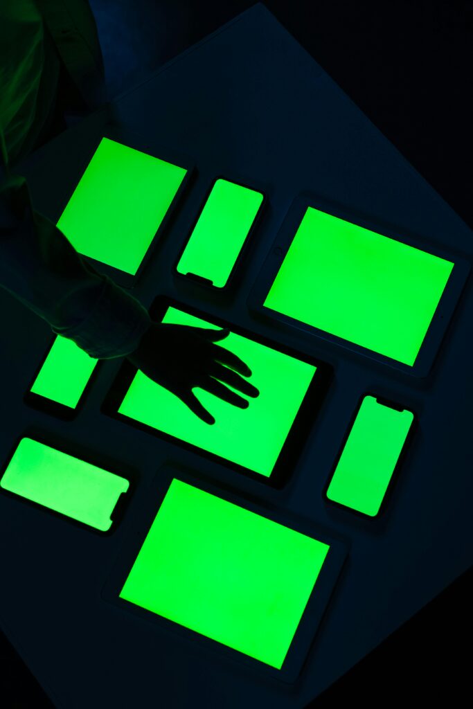 Hand reaching over various electronic devices with glowing green screens, emphasizing digital connectivity.